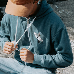A man wearing a blue zip-up hoodie with a white drawstring and a small surfboard graphic on the chest, paired with a brown hat and light blue jeans, sitting outdoors.