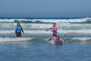 surf instructor watching a student catch a wave wearing a wetsuit and pink rash guard 